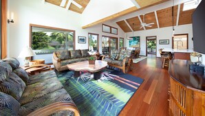 A spacious living room with a large sofa, a coffee table, and a wooden cabinet. There are multiple windows providing natural light and a view of the outdoors.