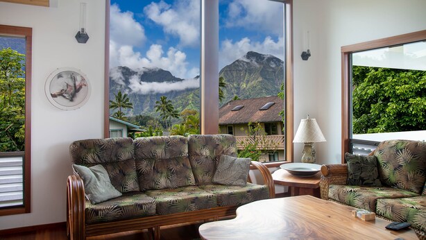 A room with a large window offering a view of mountains and clouds, a wooden sofa with a tropical print, and a wooden coffee table.