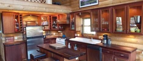 A rustic wooden cabin interior with a kitchen featuring wooden cabinets, a central island with stools, and a large window providing natural light.