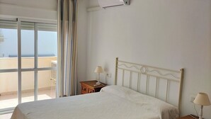 Un dormitorio con cama, ventilador de techo, unidad de aire acondicionado, mesita de noche con lámpara, cómoda de madera y vista por la ventana.