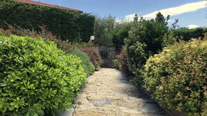 Uma trilha de pedra leva a um portão cercado por vegetação exuberante e um telhado de telhas vermelhas ao fundo.