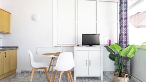 Una cocina compacta con armarios blancos, una mesa de comedor con dos sillas, un televisor sobre un mueble bajo y una planta grande.