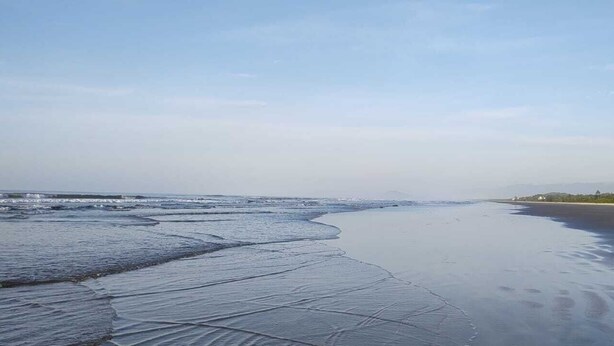 Een strand met nat zand en zachte golven, een heldere lucht en verafgelegen heuvels.
