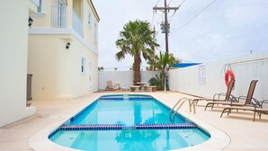 Una piscina rectangular con borde de azulejos, rodeada por una cerca blanca, una palmera y mobiliario de exterior.