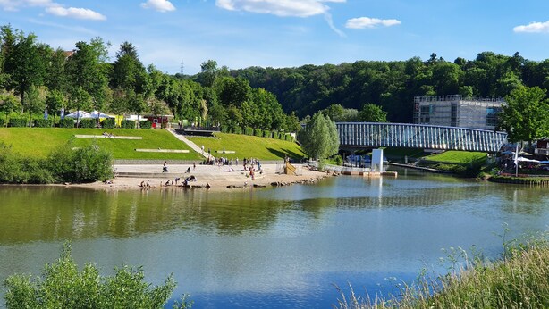 En park ved floden med en fodgængerbro, grønne arealer og et roligt vandstykke.