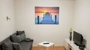En stue med en grå sofa, et hvidt sofabord, et TV på et hvidt skab og et maleri af en solnedgang på væggen.