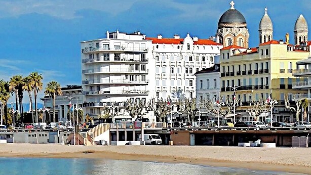 Un paysage urbain côtier avec un bâtiment imposant doté d’un dôme et de nombreuses fenêtres, une plage de sable et des palmiers.