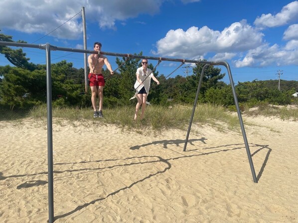 Deux personnes se trouvent sur une structure métallique, dans une zone sablonneuse, avec des arbres et un ciel dégagé.