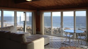 A room with a large sliding glass door leading to a beach view, a ceiling fan, a white sofa, and a small table with chairs. - Private Sandy Beach, Truly Oceanfront,Private Decks, Free Kayak, Sleeps  5 (South Yarmouth)