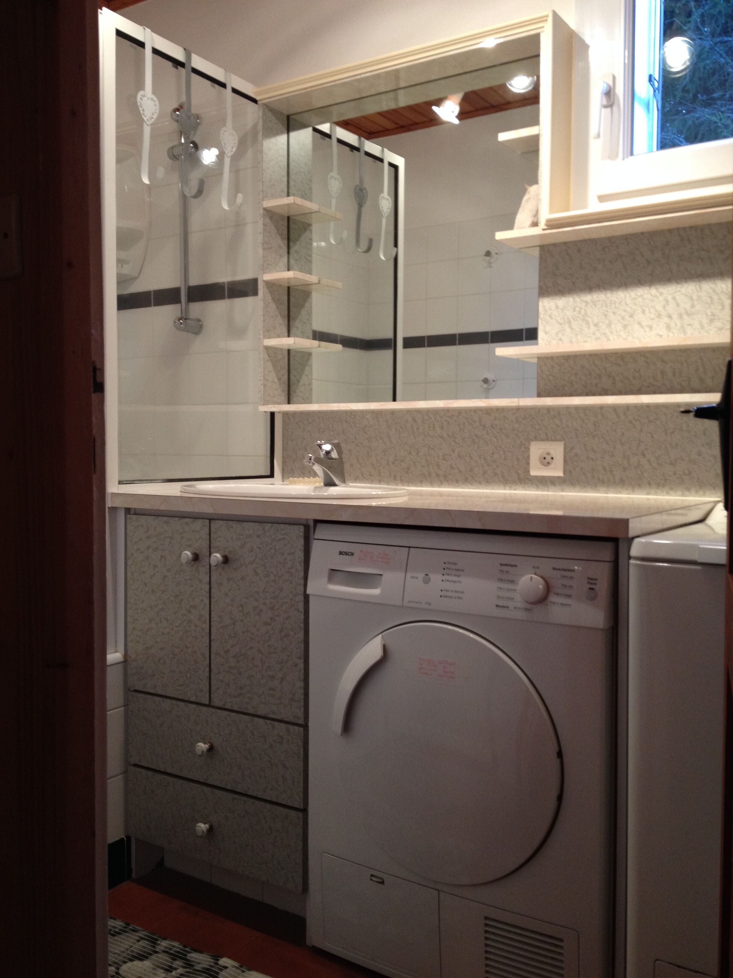Un baño compacto con lavadora Bosch, cabina de ducha de vidrio y espejo sobre el lavamanos.