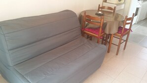 Ein graues Sofa mit Holzstühlen und einem kleinen runden Tisch in einem Zimmer mit weißen Wänden und Fliesenboden.