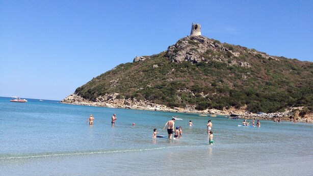 Een strand met helder water, mensen die zwemmen, en een heuvel met een toren op de achtergrond.