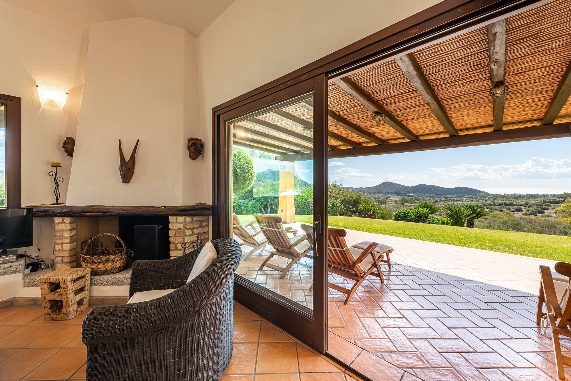 A cozy living room with a fireplace, wicker furniture, and a large sliding glass door leading to an outdoor patio with chairs and a view of the countryside.