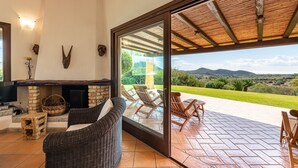 Een gezellige woonkamer met een open haard, rieten meubels en een grote schuifdeur naar een terras buiten met stoelen en uitzicht op het platteland.