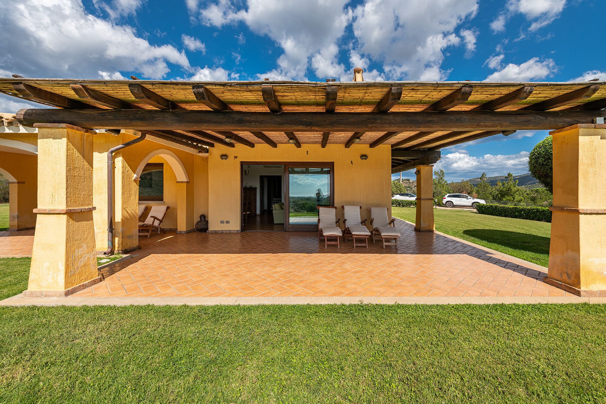 Une terrasse extérieure couverte, dotée d’un sol carrelé, d’une pergola et offrant une vue sur une maison à travers une porte vitrée.
