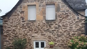 Il s’agit d’un bâtiment en pierre, doté d’un toit à pignon, de deux fenêtres avec volets et d’une petite porte. Des plantes en pot sont disposées devant le bâtiment.