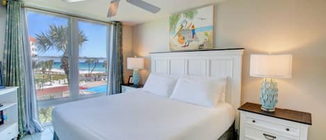 A hotel room with a large bed, a ceiling fan, a nightstand with a lamp, and a painting of a beach scene.