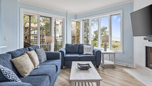 Una sala de estar con un sofá azul, una mesa de centro y un televisor. Se ven ventanas grandes con vistas a edificios y árboles.