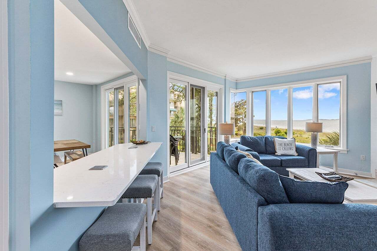 A modern kitchen with a white countertop and bar seating, a blue sofa with a beach-themed pillow, and a view of the beach through large windows.