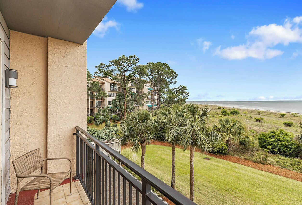 A balcony with a bench, overlooking a grassy area with palm trees and a view of a building complex and the ocean.