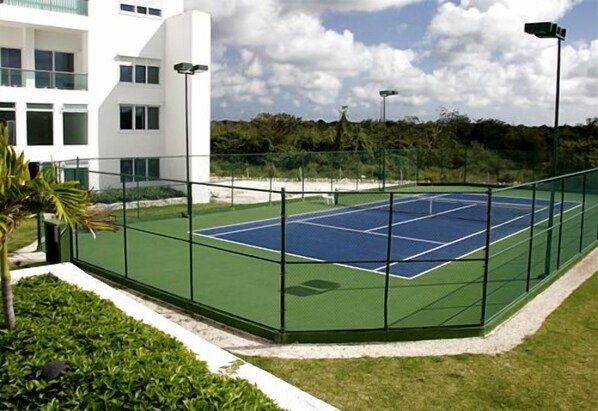 Una cancha de tenis al aire libre con valla verde, rodeada por un edificio blanco y abundante vegetación.