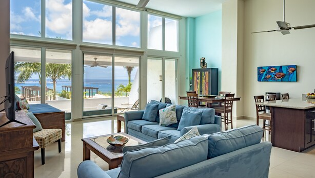 Amplia sala de estar con un gran ventanal con vista a una playa, un comedor y una cocina con barra.