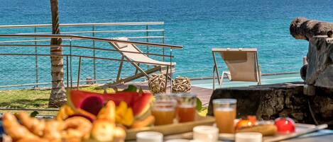 A breakfast setup with croissants, fruits, and beverages on a terrace overlooking the sea.