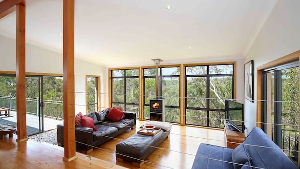 A spacious living room with a large black leather sofa, a blue armchair, and a wooden coffee table. A wood-burning stove is visible through a large window, and the room has a high ceiling with exposed wooden beams. - Postcards From The Edge (Blackheath)