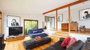 A modern living room with a blue sofa, a black leather couch, and a wooden coffee table. There is a television on the left and a wooden cabinet on the right. - Postcards From The Edge (Blackheath)