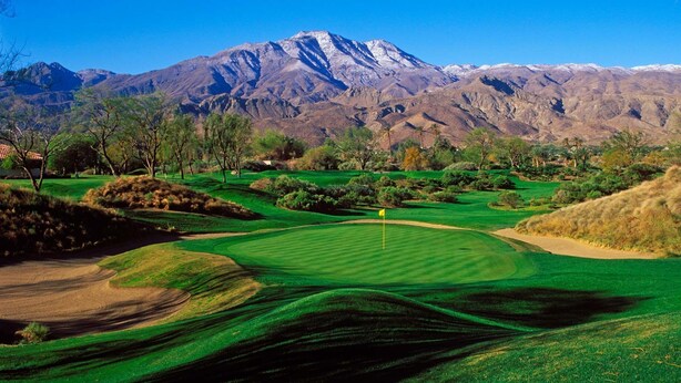 Un campo de golf con una bandera en el centro, rodeado de césped cuidado y hoyos de arena, con una cadena montañosa al fondo.
