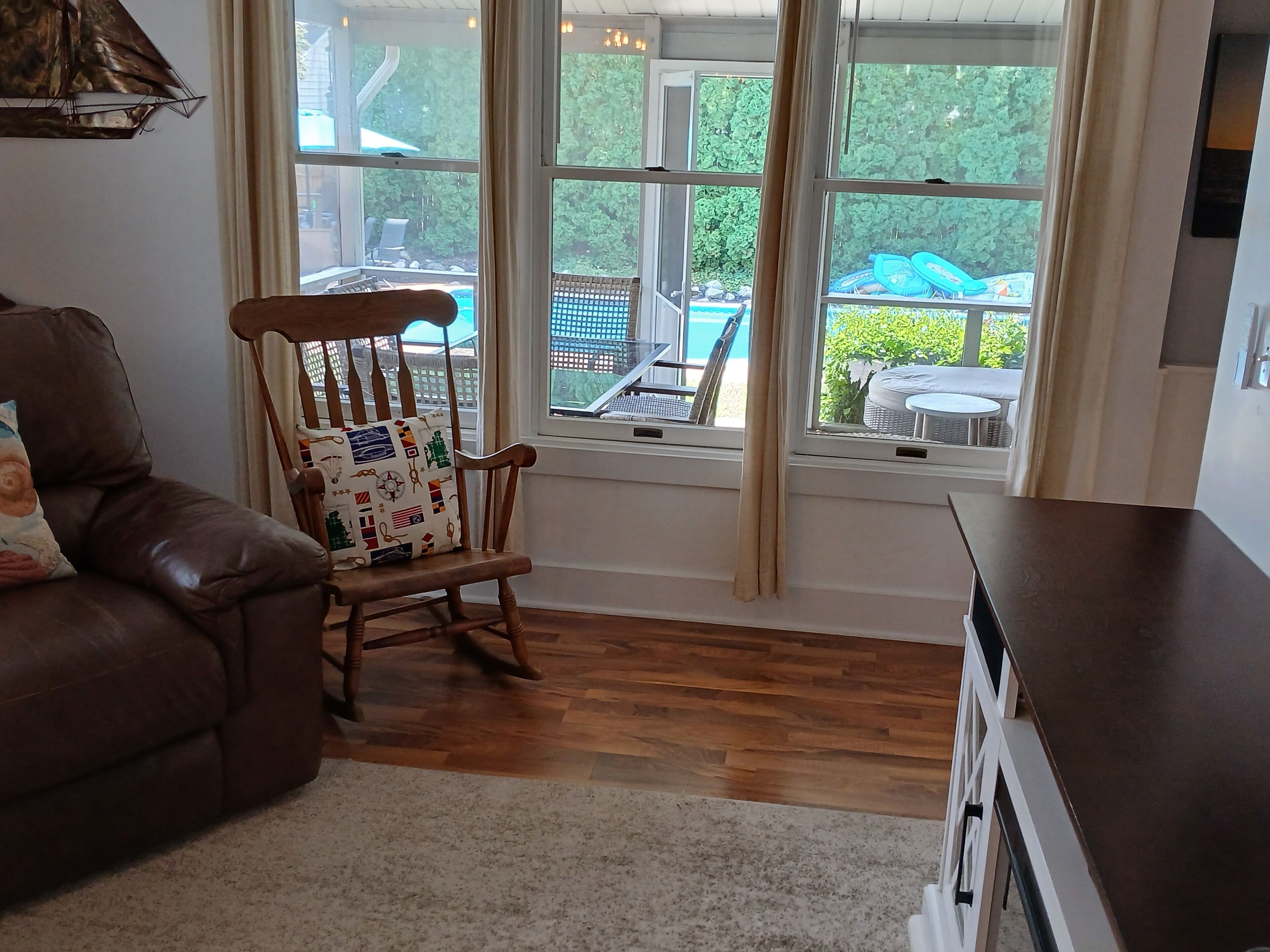 A living room with a brown leather sofa, a wooden rocking chair with a patterned cushion, a white rug, and a large window with a view of outdoor furniture and greenery.