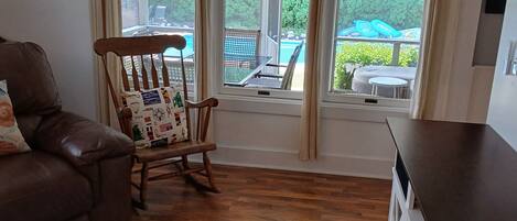 A living room with a brown leather sofa, a wooden rocking chair with a patterned cushion, a white rug, and a large window with a view of outdoor furniture and greenery.