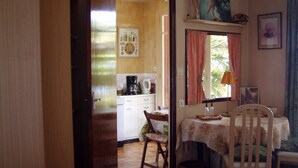 Una habitación con una mesa de comedor, sillas, un ventanal con cortinas, una zona de cocina al fondo y una puerta que da a otra habitación.