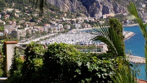 Un paisaje urbano costero con edificios densos, un puerto deportivo con numerosos barcos y una ladera rocosa al fondo.
