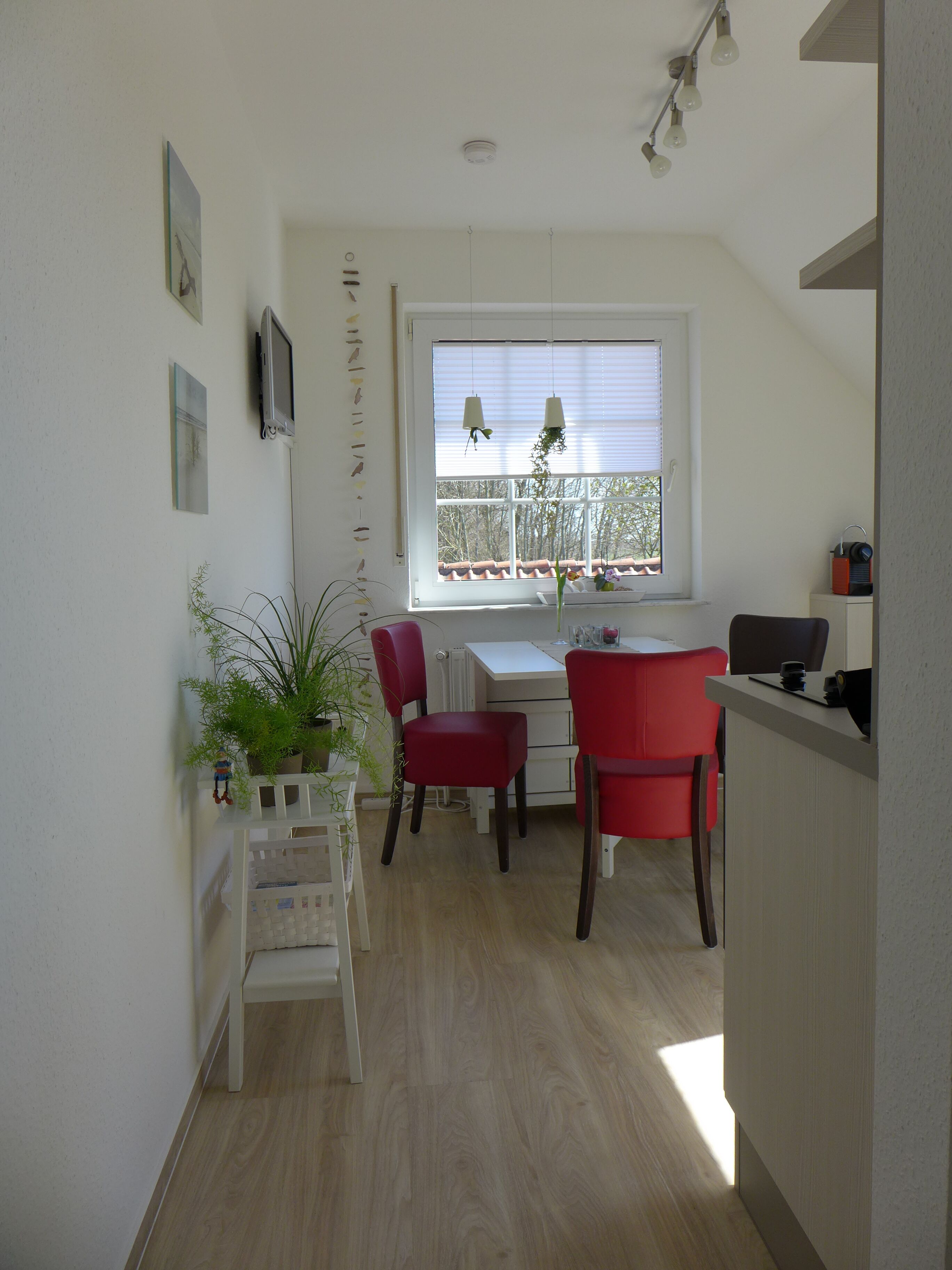 A small, bright room with a white table, two red chairs, a potted plant, and a window with blinds.