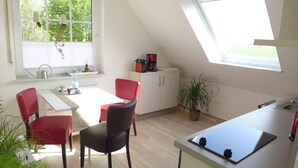 A modern kitchen with a dining area, featuring a white table, red chairs, a coffee machine, and a potted plant. - Bright, friendly top floor apartment with WLAN, 15 min. walk to the beach. (Norden)