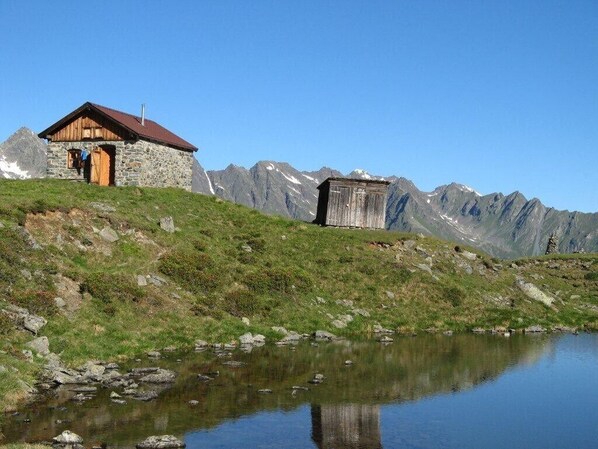 A stone building with a wooden door and a red roof, situated on a grassy hill with a mountain backdrop and a calm lake in the foreground.