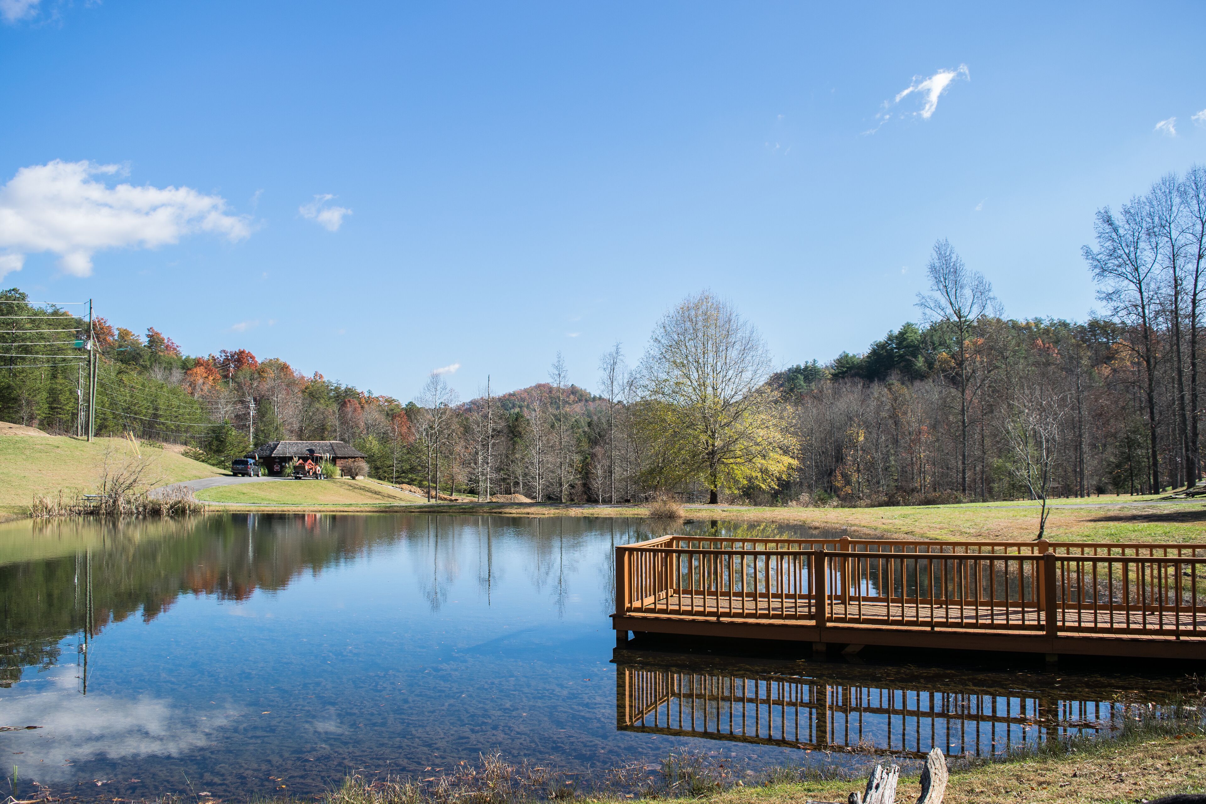 A wooden deck over a calm lake with a reflection, surrounded by trees and a small building.