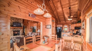 Un acogedor interior de cabaña de troncos con chimenea de piedra, mesa de comedor de madera y zona de cocina.