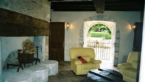 Een gezellige kamer met een stenen openhaard, een gele fauteuil en uitzicht op een tuin door een rondboogvenster.