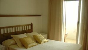 Un dormitorio con cabecera de madera, cama con ropa blanca y dos almohadas, mesita de noche y vista al exterior a través de una ventana con cortinas.
