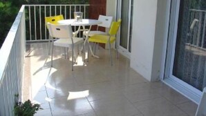Un balcón con una mesa y sillas blancas, un ventanal de vidrio y vista a follaje.