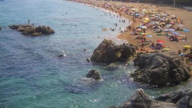 Una playa con afloramientos rocosos y un horizonte urbano al fondo.