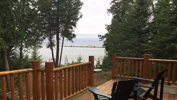 A wooden deck with a railing overlooking a lake, two chairs, and a small table. - Gorgeous Rustic Northern Michigan Island Getaway (St. James)