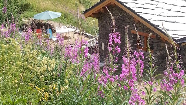 A stone building with a sloped roof, surrounded by lush greenery and purple flowers. - Chalet on the slopes in Valmeinier 1800 (Valmeinier)