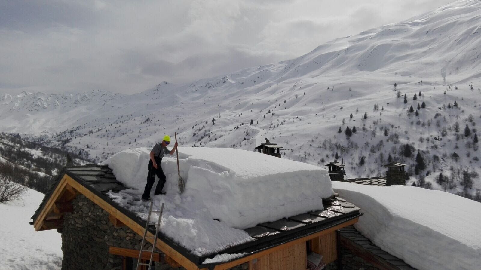 A person shoveling snow from a roof of a stone building with a wooden structure on a snowy mountain slope.