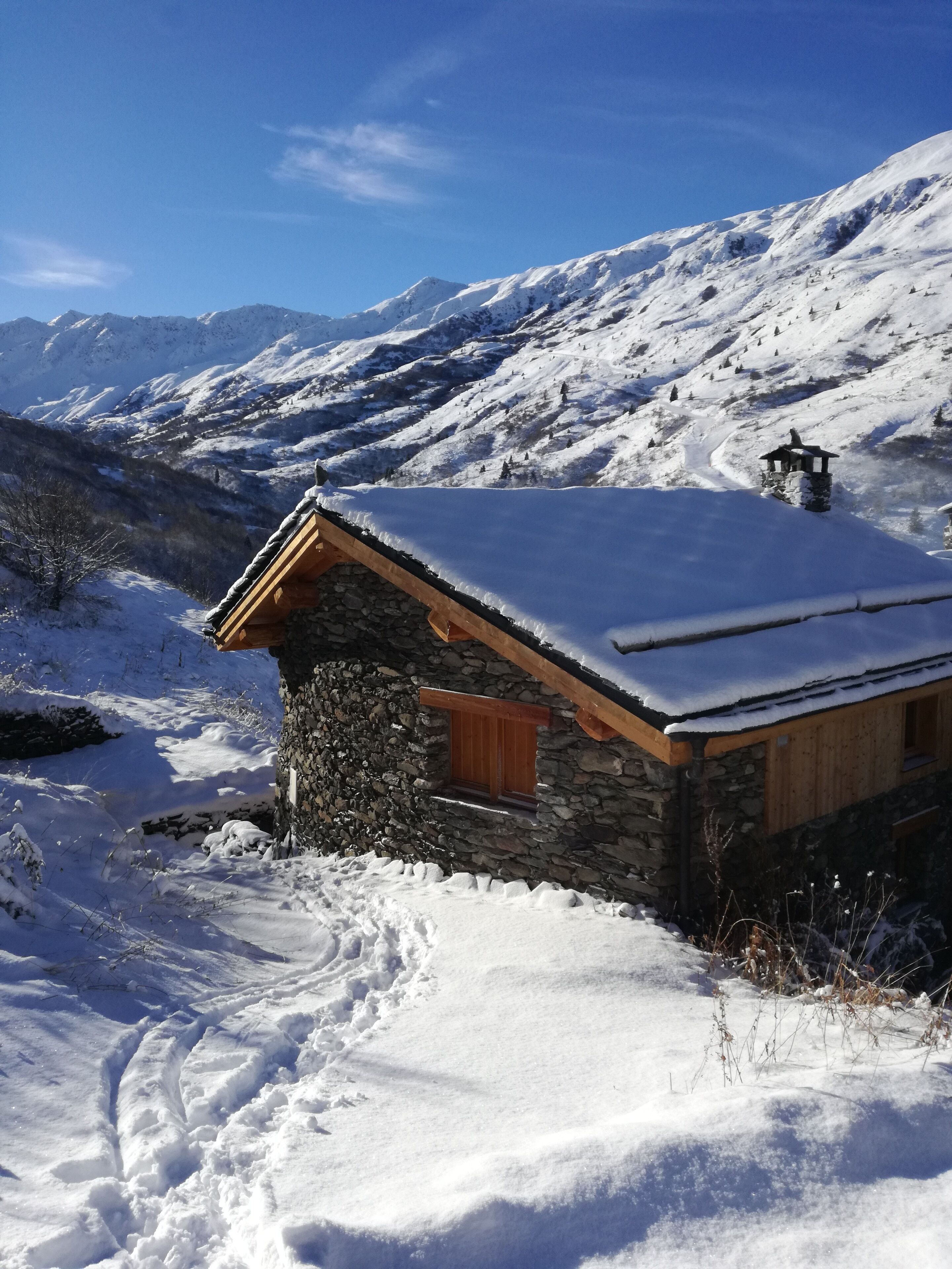 Een stenen huisje met een houten dak en een klein raam, omgeven door besneeuwde bergen en een heldere lucht.
