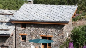 A stone building with a sloped roof, wooden windows, and a small outdoor seating area with a green umbrella. - Chalet on the slopes in Valmeinier 1800 (Valmeinier)