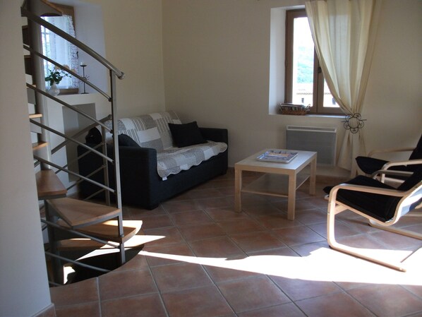 A living room with a sofa, a coffee table, a chair, a staircase, and a window with curtains. - BASTIDON, POOL, 3000 M2 OF LAND, 2-4 PERS, APT, Luberon (Apt)