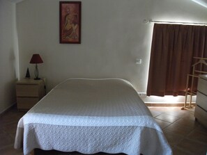 Une chambre à coucher avec un lit, une table de chevet, une lampe, un tableau et une fenêtre avec des rideaux.